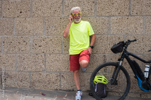 Old caucasian man with white hair and beard, talks at his cell phone near his bicycle. Brick wall as a background. Dressed in a colorful way, with yellow sunglasses. One people only