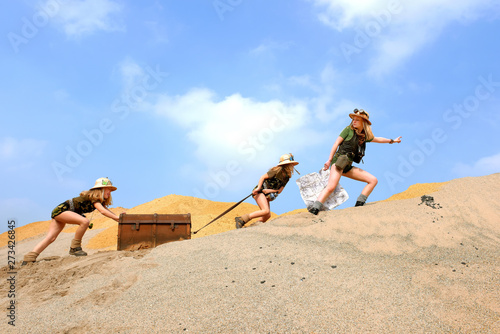 Young twin sisters and an adult girl dress up as explorers. They pretend to be hauling an old luggage chest up a sand dune hill. The girls are dressed in khaki safari clothes and wear jungle hats.