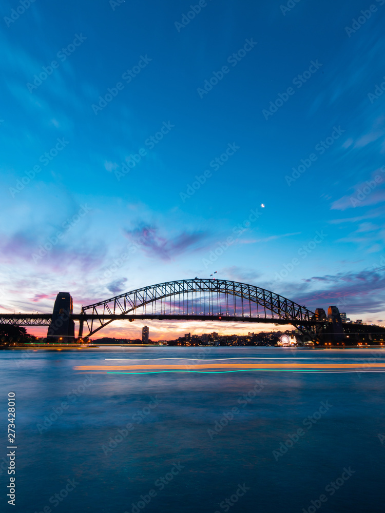 Boat trail passing through Sydney Harbour Bridge at dusk.