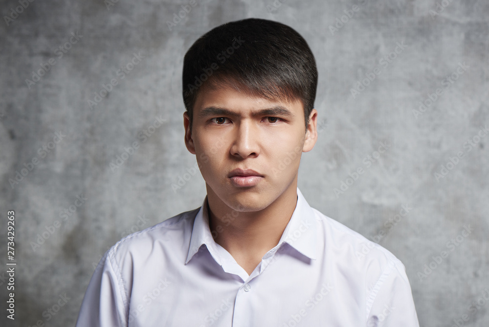 Serious asian student with evil face and frowning eyebrows on gray background. Emotional Kazakh looks with displeasure at camera Serious asian student with evil face and frowning eyebrows on gray background. Emotional Kazakh looks with displeasure at camera