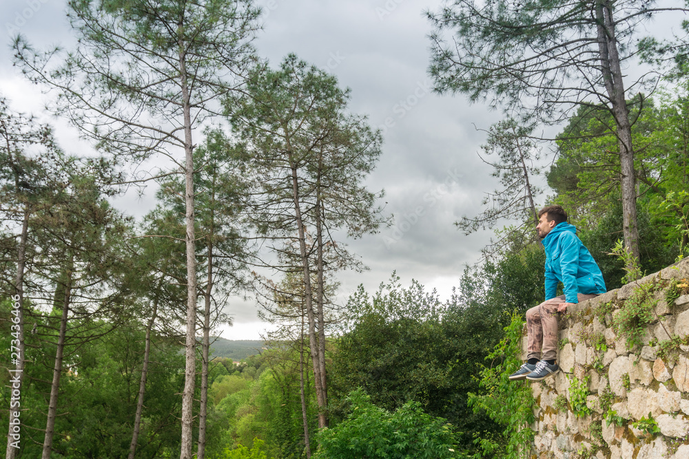Young man sitting alone on stones wall and looking at green nature view with forest on background. Girona, Catalonia, Spain. Man in nature concept. Young man sitting alone on stones wall and looking at green nature view with forest on background. Girona, Catalonia, Spain. Man in nature concept.
