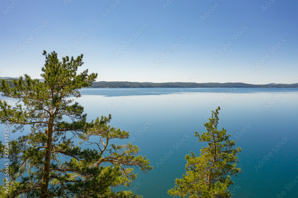 Obraz premium Aerial footage of picturesque summer nature; drone flying backward to forest thicket growing on rocky shore of mountain lake; clear skyline with hills in background; protected freshwater environment