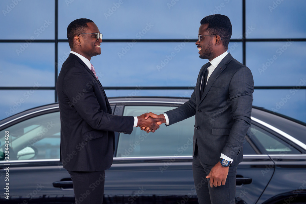 two successful african american business men well dressed partners standing next to the luxury car on the background of the offices windows two successful african american business men well dressed partners standing next to the luxury car on the background of the offices windows