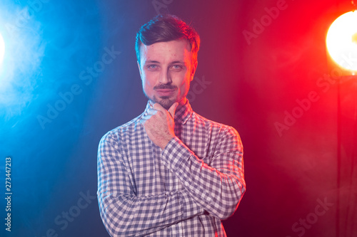 Carming young smiling hipster man is posing against the background of red and blue studio lighting. Concept of pensive man.