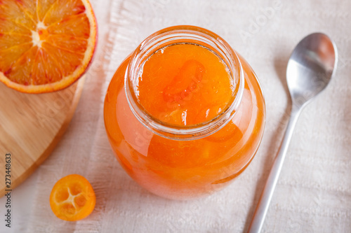 Orange and kumquat jam in a glass jar with fresh fruits on a white linen tablecloth.