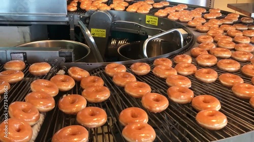Freshly baked with a warm sugar glaze donuts going round on a conveyor belt waiting to be boxed by the baker. Filmed in 4k at cinematic 24 fps.