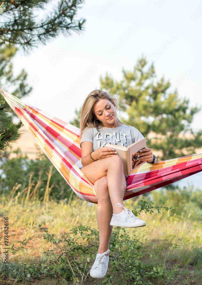 Obraz premium young attractive caucasian woman reading book in hammock in the forest, lake on the background