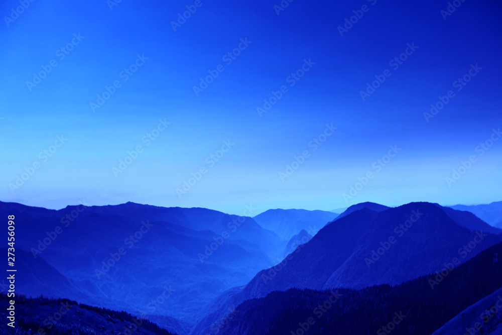 Fototapeta premium The outlines of mountains and hills through the morning atmospheric fog in neon blue color. A picturesque place for trekking. Fantasy postcard, copyspace.