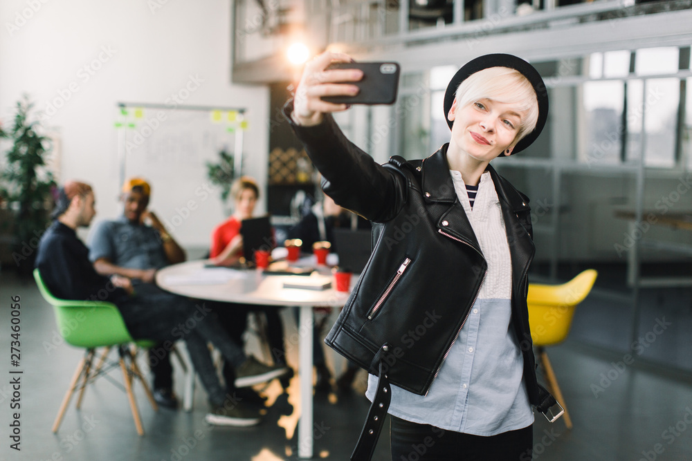 Cheerful girl in black hat and leather jacket making video call via smartphone. Young woman waving hand and smiling at phone screen. Video call concept Cheerful girl in black hat and leather jacket making video call via smartphone. Young woman waving hand and smiling at phone screen. Video call concept