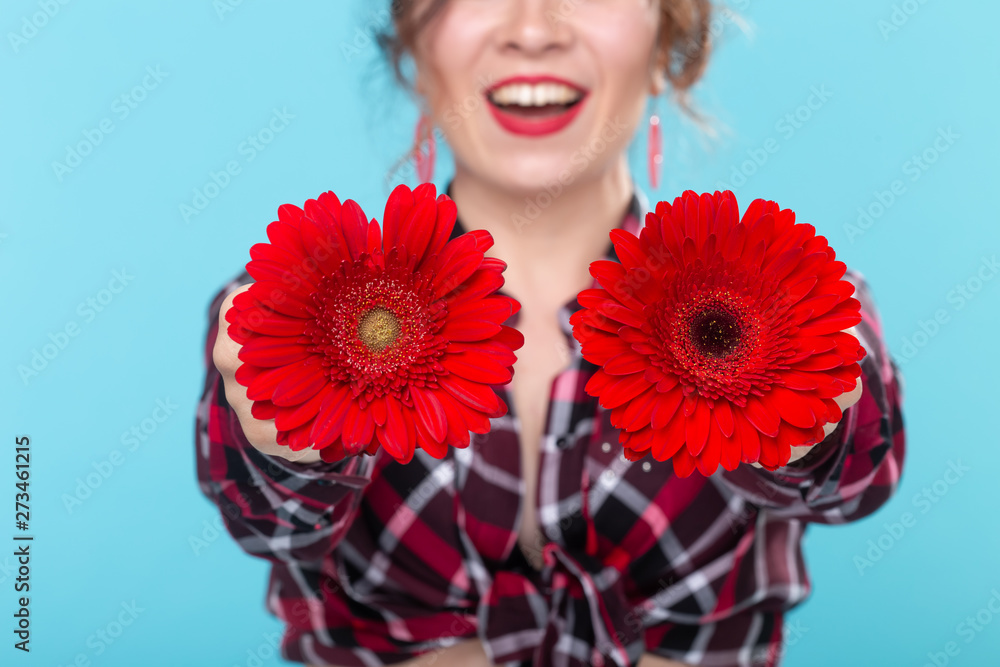 Close-up of an unidentified young smiling woman holding two red flowers with different cores posing in front of a blue background in front of her. Concept distinction nuances Close-up of an unidentified young smiling woman holding two red flowers with different cores posing in front of a blue background in front of her. Concept distinction nuances