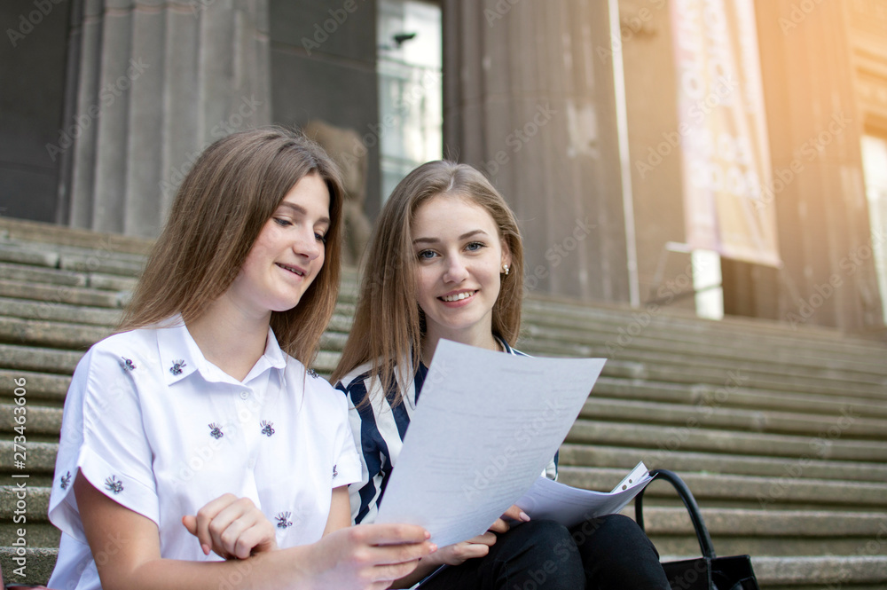 Two beautiful students sit on the steps near the school, hold paper in their hands and learn lessons, they communicate at the university during the break Two beautiful students sit on the steps near the school, hold paper in their hands and learn lessons, they communicate at the university during the break