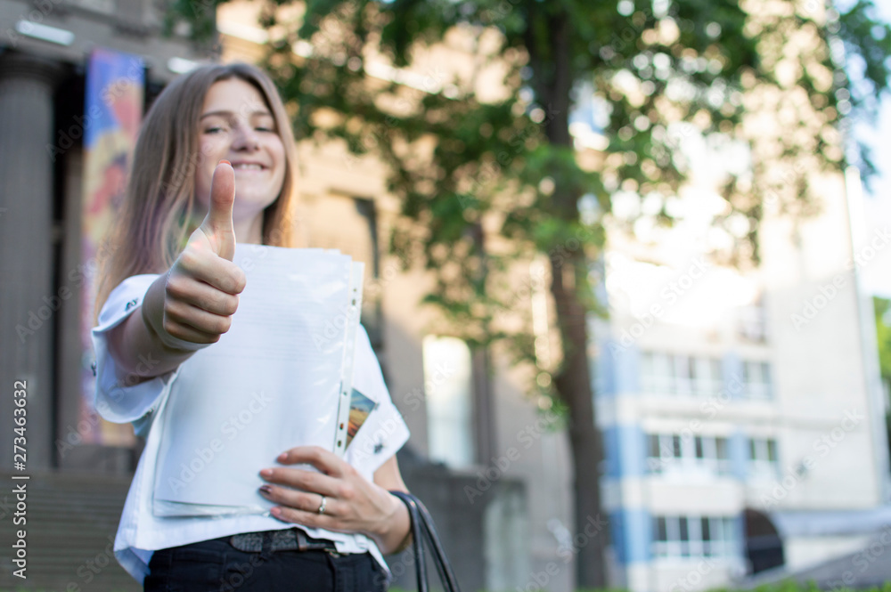 young student standing near the university, holding a paper in her hands smiling and showing like against the background of the college, she goes to school young student standing near the university, holding a paper in her hands smiling and showing like against the background of the college, she goes to school