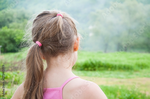 a girl in a vest with a haircut is standing in a meadow. Back view. Hair laid in ponytail