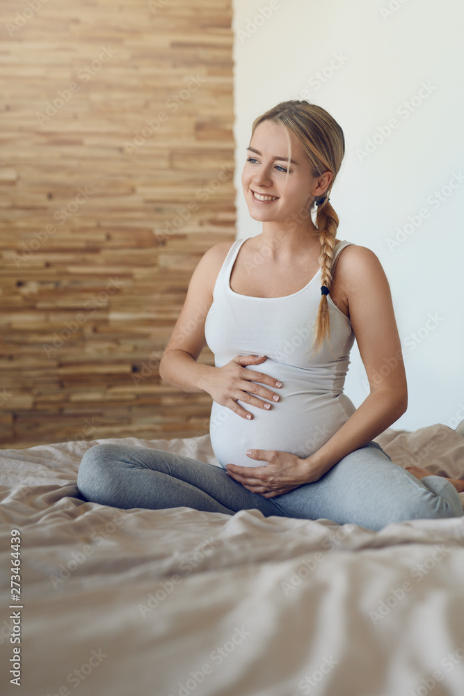 Happy young pregnant woman bonding with her unborn child sitting on a bed cradling her baby bump with her hands looking sideways with a tender smile of love Happy young pregnant woman bonding with her unborn child sitting on a bed cradling her baby bump with her hands looking sideways with a tender smile of love