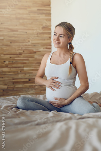 Happy young pregnant woman bonding with her unborn child sitting on a bed cradling her baby bump with her hands looking sideways with a tender smile of love