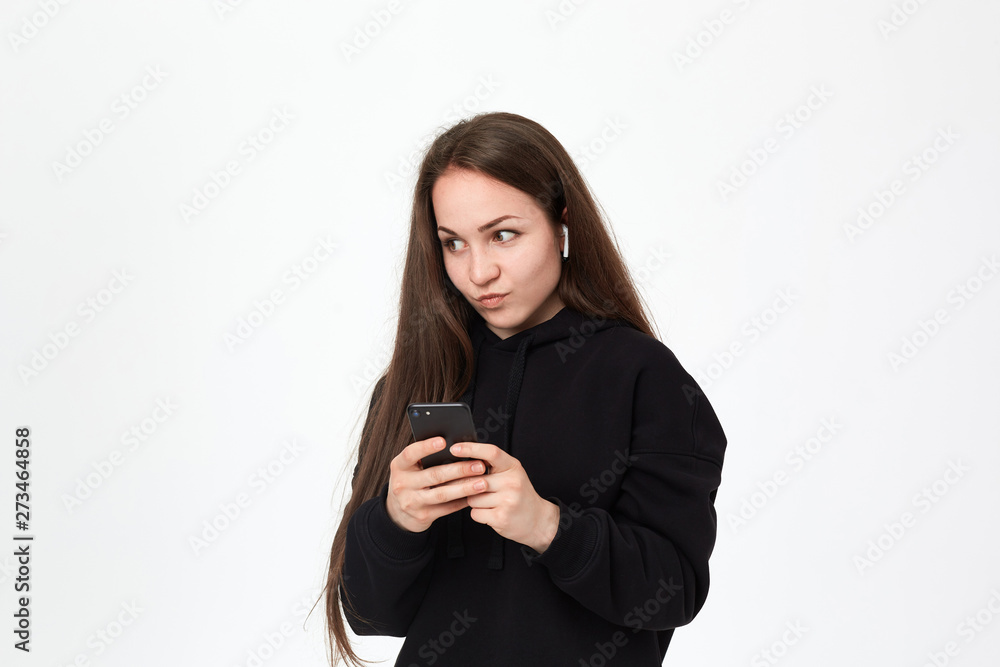 Studio shot of a beautiful young brunette woman with wireless headset and phone looking intrigued at the right side while standing over white background. Studio shot of a beautiful young brunette woman with wireless headset and phone looking intrigued at the right side while standing over white background.