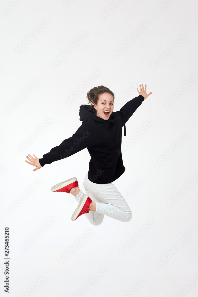 Young pretty brunette girl in black hoodie, white pants and red sneakers jumping isolated on white background.
