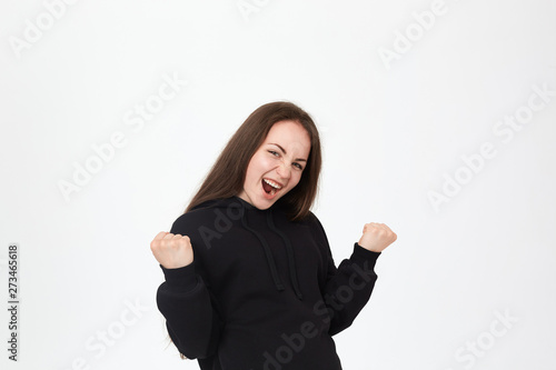 Studio shot of a successful pretty young brunette girl standing over white background. Model shows positive gesture of success at the camera.