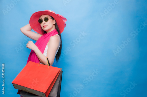 Portrait of an excited beautiful girl wearing dress (white T-Shirt), pink hat and sunglasses holding shopping bags isolated over blue background.