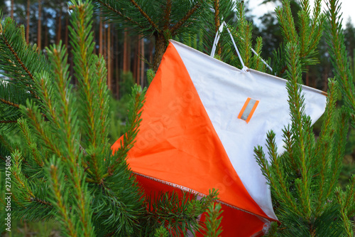Orange check point prism for orienteering or rogaining competition hanging on green spruce tree branches outdoors in summer day. Sport equipment concept.