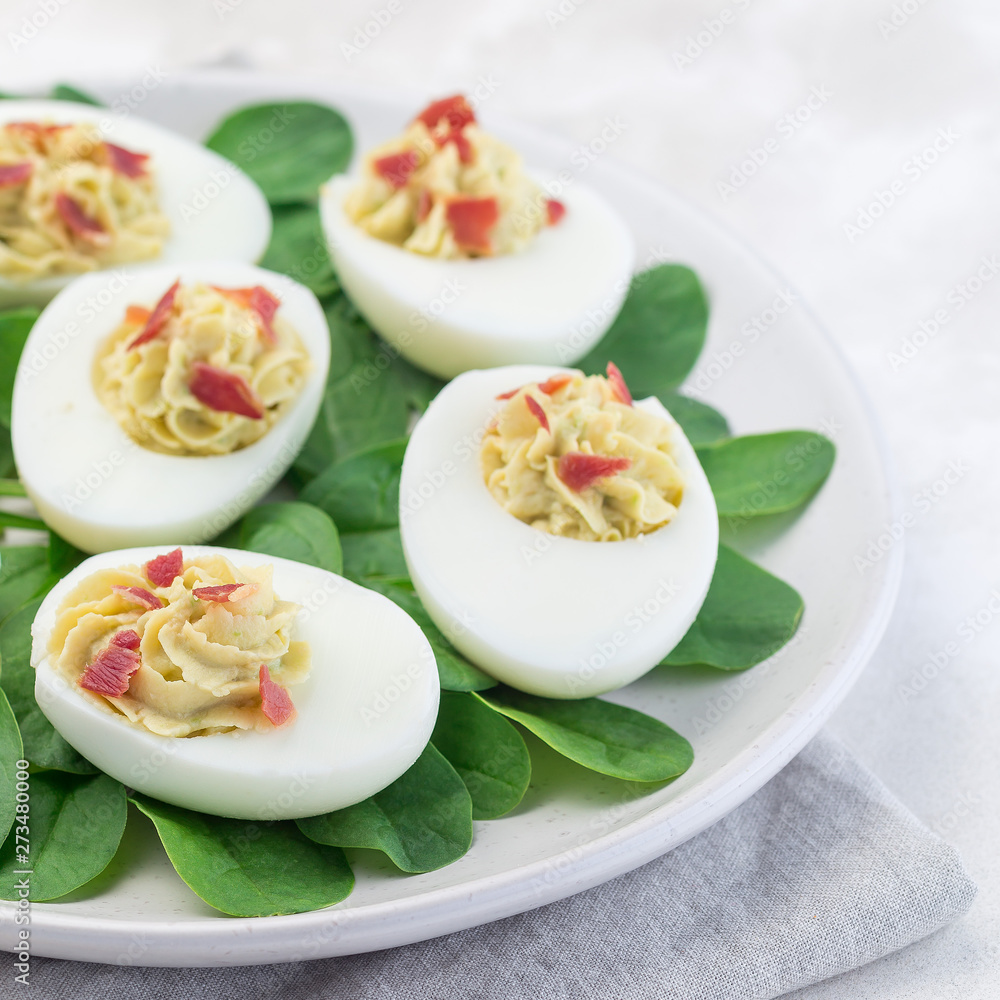 Deviled eggs stuffed with avocado, egg yolk and mayonnaise filling, garnished with bacon on spinach leaves, square format Deviled eggs stuffed with avocado, egg yolk and mayonnaise filling, garnished with bacon on spinach leaves, square format