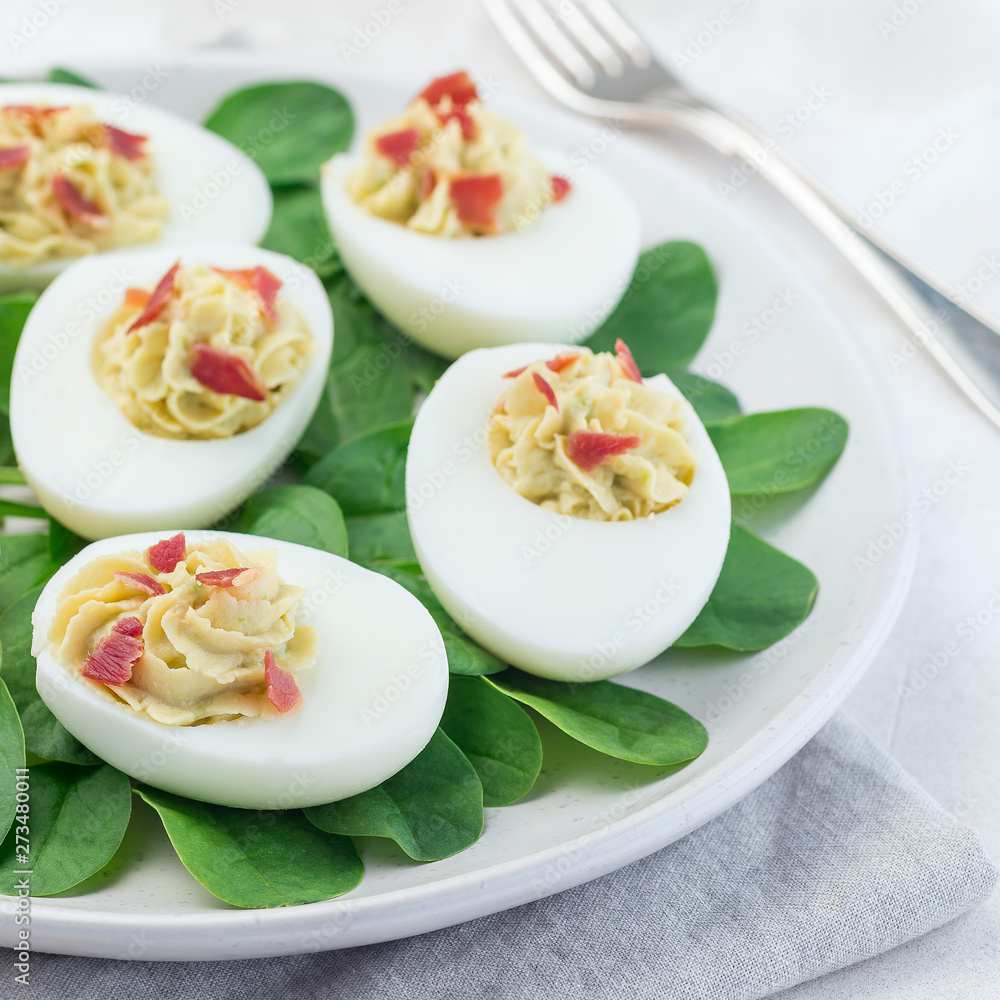 Deviled eggs stuffed with avocado, egg yolk and mayonnaise filling, garnished with bacon on spinach leaves, square Deviled eggs stuffed with avocado, egg yolk and mayonnaise filling, garnished with bacon on spinach leaves, square