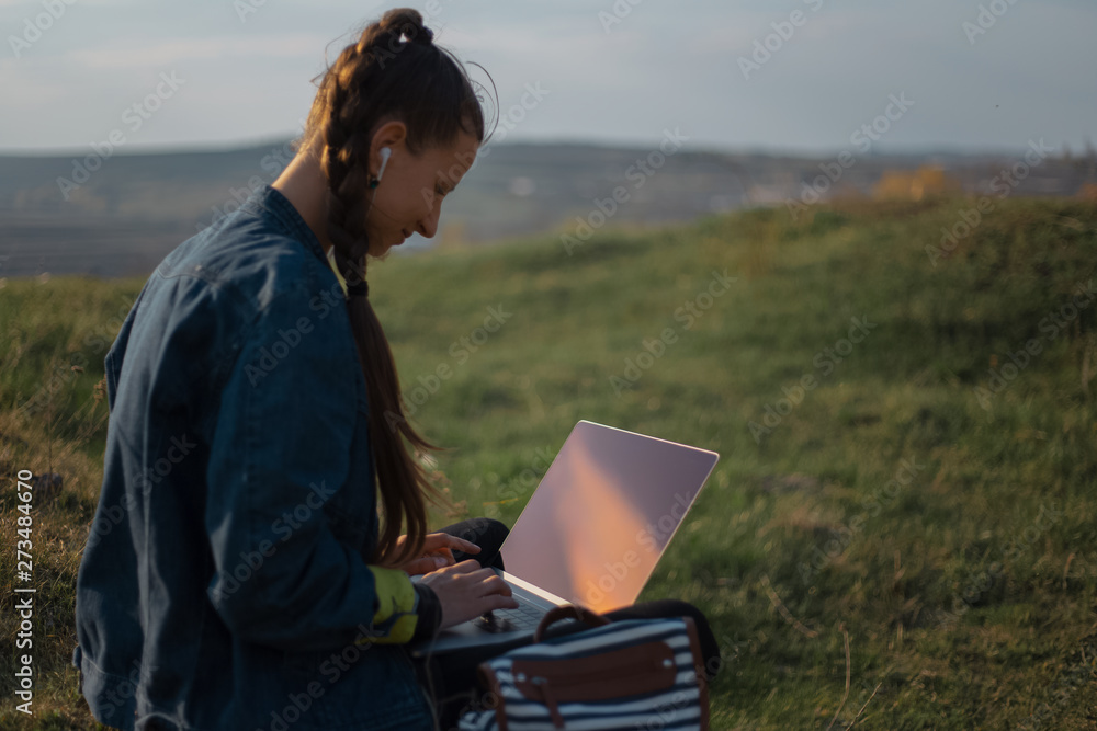 young girl working at laptop with earphones in the fields 