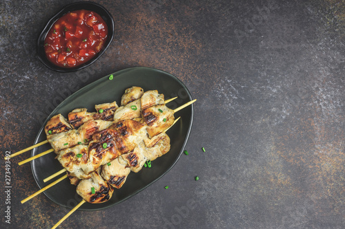 Grilled chicken skewers with green onion and salsa sauce in a plate on a dark background. Top view.