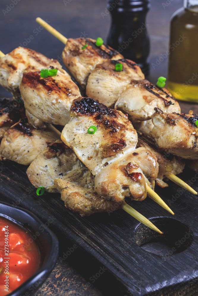 Grilled chicken skewers with green onion and salsa sauce in a cutting board on a dark background. Selective focus