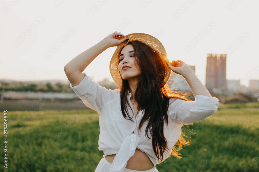 Obraz premium Portrait of beautiful carefree long hair asian girl in white clothes and straw hat enjoys life in field at sunset. Sensitivity to nature concept
