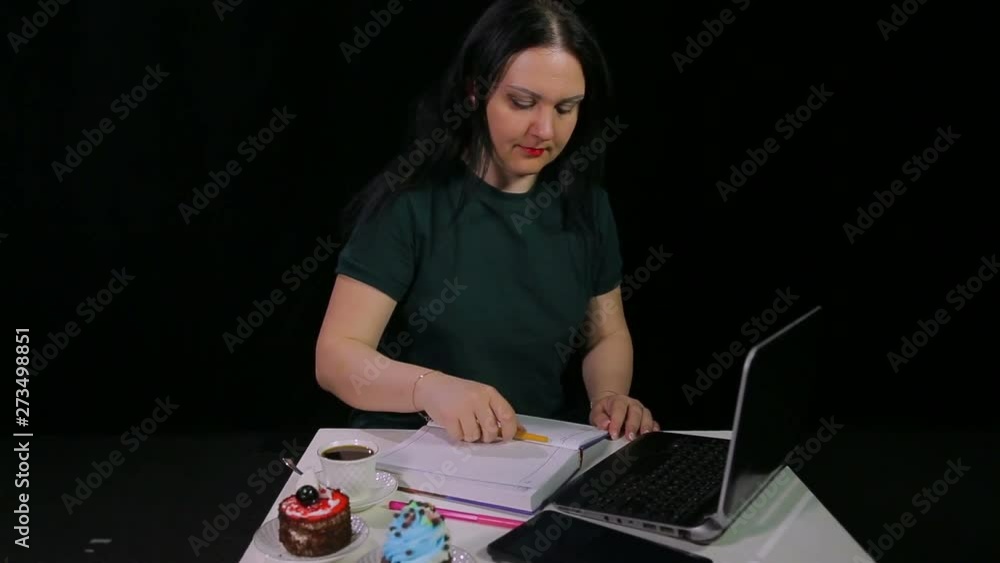 A woman in a cafe drinks coffee from a white cup on a black background and writes in a diary while looking into a computer A woman in a cafe drinks coffee from a white cup on a black background and writes in a diary while looking into a computer