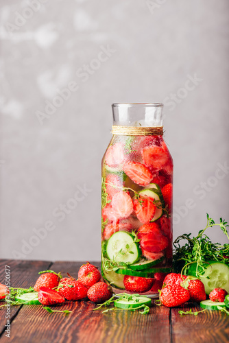 Bottle of Infused Water with Fresh Strawberry, Sliced Cucumber and Springs of Thyme. Ingredients Scattered on Wooden Table. Copy Space and Vertical Orientation.