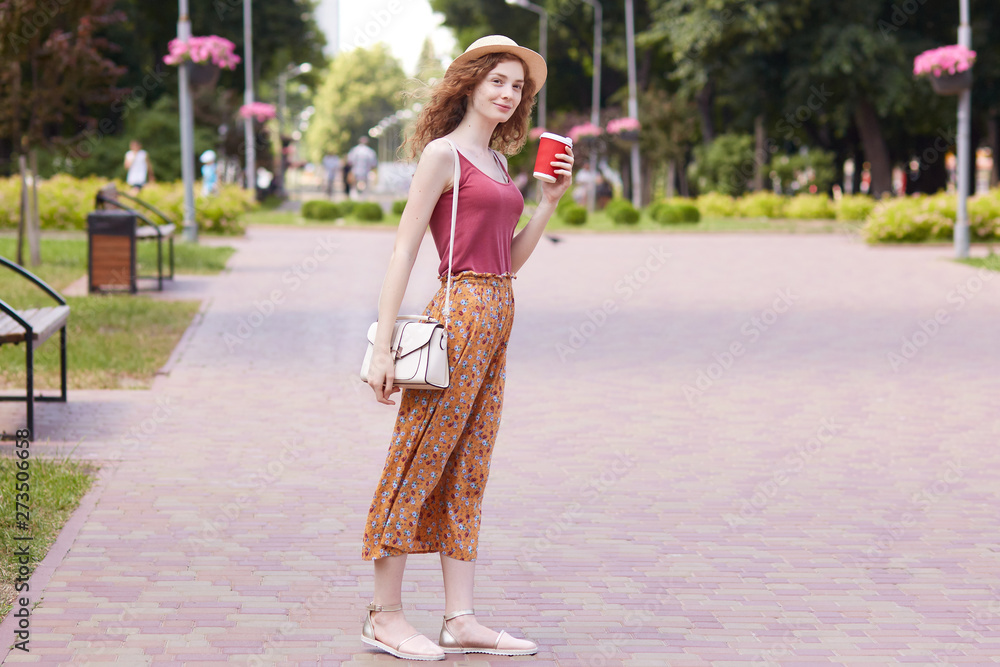 Full length portrait of slender elegant young female walking through recreation zone in native town, wearing hat, red top and summer skirt, having white trendy bag on shoulder, holding takeaway drink.