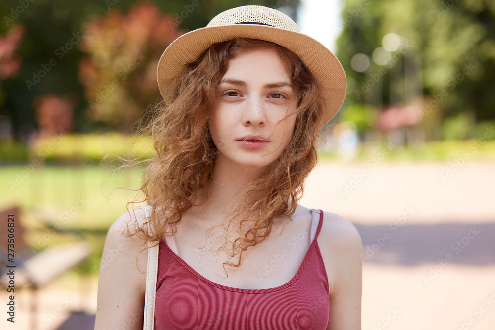 Outdoor shot of thoughtful attentive cute girl having mysterious facial expression, having walk at local recreation zone, being pensive, posing with fair curly hair, wearing casual clothes. Mood.