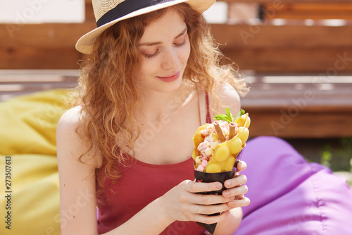 Horizontal shot of cute young beautiful woman in hat and t shirt, being in city park with ice cream, having rest after studying and having fun with friends, spends time in open air. People concept photo