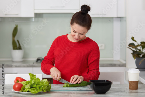 Indoor shot of pregnant woman cooking at home, doing fresh green salad for dinner, eating many different vegetables during pregnancy, healthy pregnancy concept, lady wearing red casual sweater.