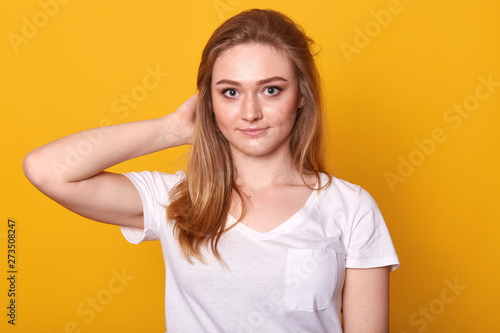 Closeup portrait of positive energetic young lady touching her hair with one hand, looking directly at camera, having pleasant facial expression, standing isolated over yellow background, being calm.