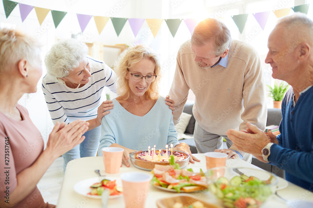 Smiling excited mature birthday woman with curly hair sitting at table and thinking about wish while preparing to blow candles on sweet cake Smiling excited mature birthday woman with curly hair sitting at table and thinking about wish while preparing to blow candles on sweet cake