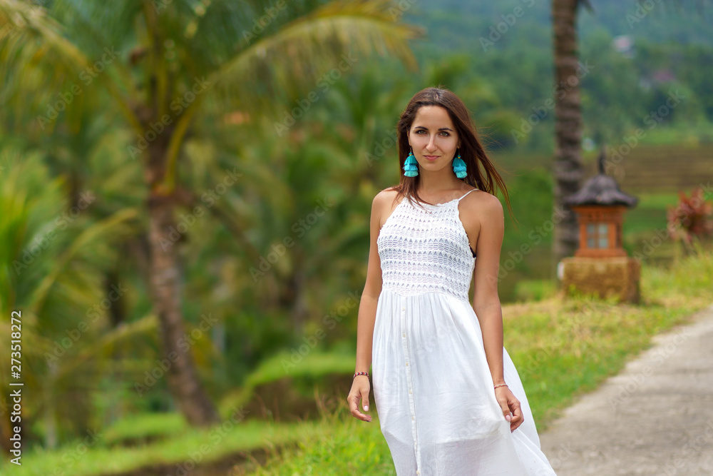 Young tanned woman in white dress posing standing on the road. In the background are palm trees and other tropical vegetation. Close up Young tanned woman in white dress posing standing on the road. In the background are palm trees and other tropical vegetation. Close up