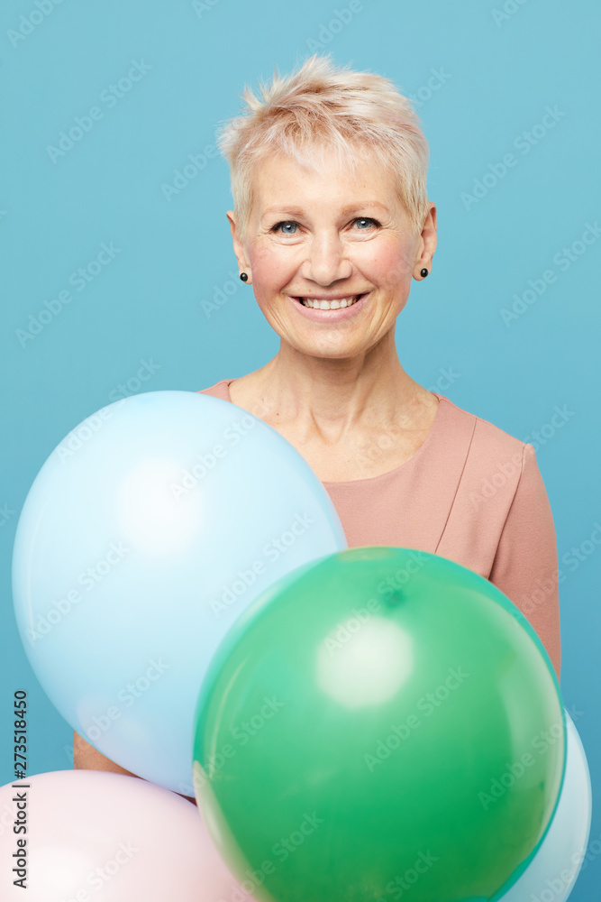 Portrait of jolly beautiful mature lady with short hair standing against blue background and holding colorful balloons Portrait of jolly beautiful mature lady with short hair standing against blue background and holding colorful balloons