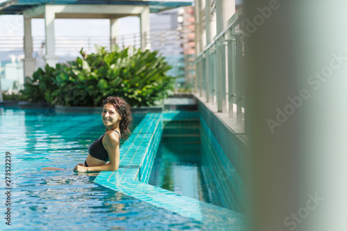 Portrait of a sexy smiling caucasian woman in a swimsuit relaxing in a rooftop pool with green bushes and city views. Asia weekend