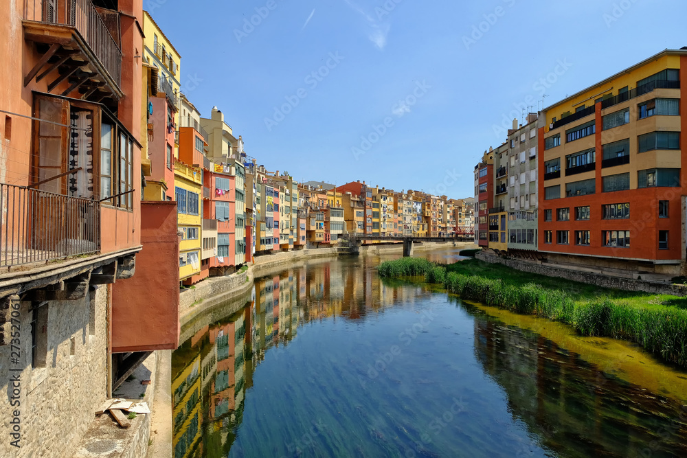 Colorful yellow and orange houses and Eiffel Bridge, Old fish stalls, reflected in water river Onyar, in Girona, Catalonia, Spain. Church of Sant Feliu at background.