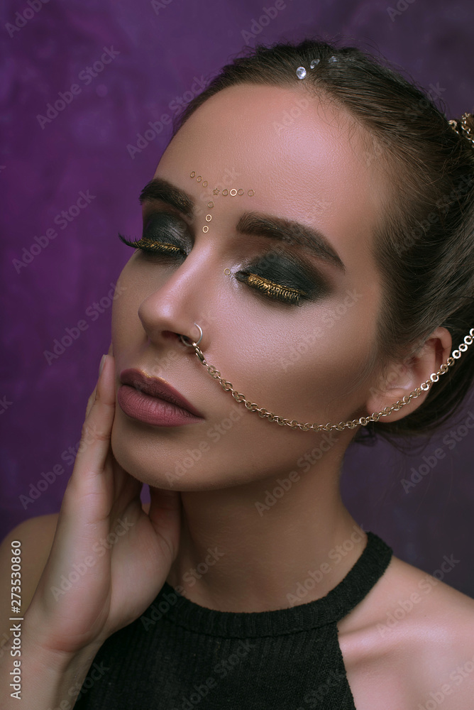 Girl with cool fashionable make-up posing in studio,matte lips, smokey gold eyes, piercing chain from nose over purple dark background with magic glow. Holiday disco celebration Girl with cool fashionable make-up posing in studio,matte lips, smokey gold eyes, piercing chain from nose over purple dark background with magic glow. Holiday disco celebration