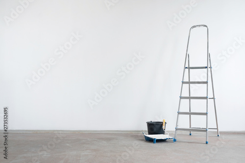 Ladder and bucket with paint roller on a white wall background with copy space. Under construction concept background.