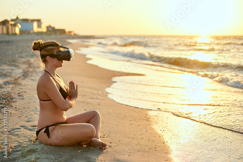 Woman in VR glasses doing yoga padmasana on ocean beach in the morning. Girl using virtual reality for deep immersion to feel herself at the seaside concept. Yoga online internet lessons with coach. © artiemedvedev