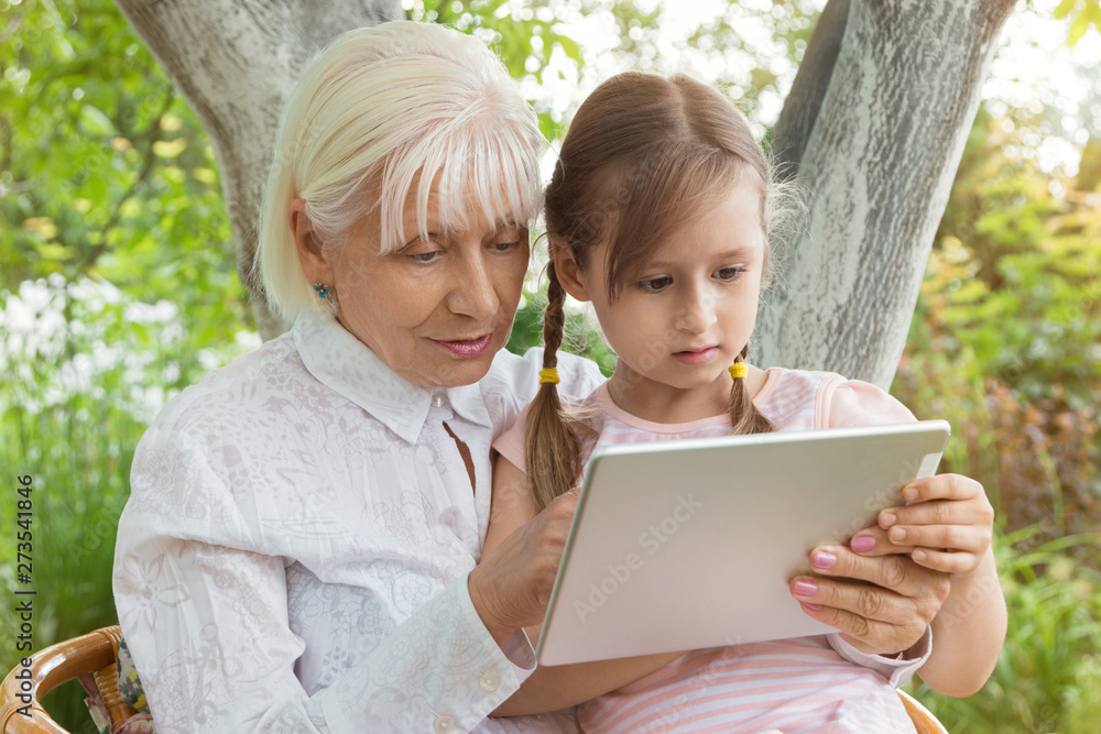 Grandmother with her granddaughter studying a book on a tablet, work on the tablet. Granny at the age of 70 years old with great granddaughter in her arms. Concept. Relationship in the family. Grandmother with her granddaughter studying a book on a tablet, work on the tablet. Granny at the age of 70 years old with great granddaughter in her arms. Concept. Relationship in the family.