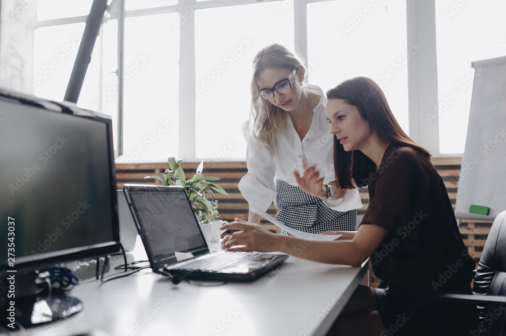 Fototapeta premium Two young beautiful women dressed according to office dreskod are working together with laptop at the desk in a light modern office