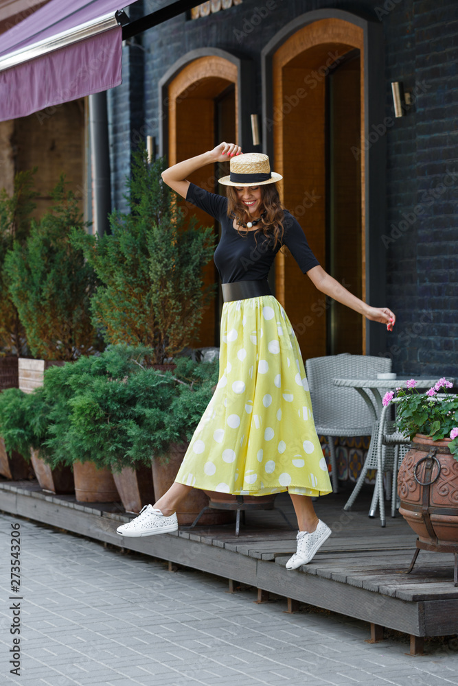 Fototapeta premium Stylish brunette girl dressed in black blouse and long yellow in white peas skirt walks in the city street on a sunny day