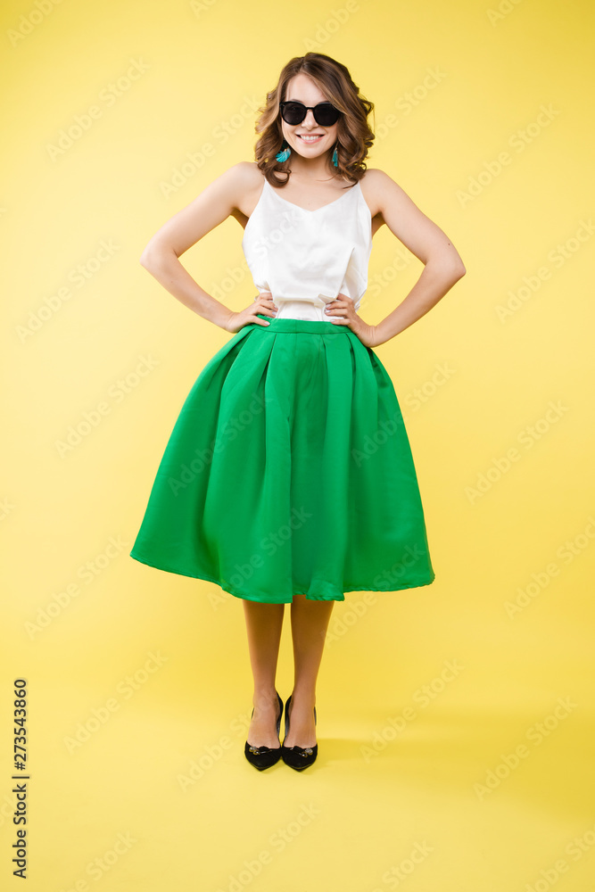 Summer outfit on orange background. Stylish and trendy beautiful young girl with wavy middle-length hair in white top and green bottom wearing trendy earrings and black sunglasses. She is smiling over Summer outfit on orange background. Stylish and trendy beautiful young girl with wavy middle-length hair in white top and green bottom wearing trendy earrings and black sunglasses. She is smiling over
