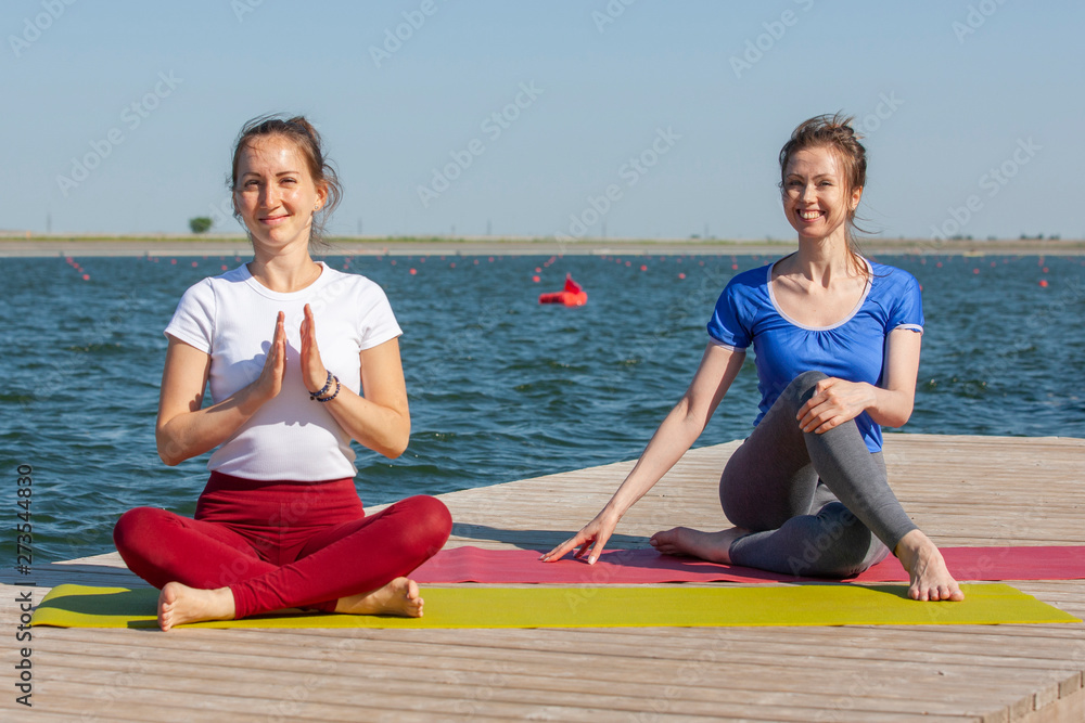 Two young women doing yoga at nature. Fitness, sport, yoga and healthy lifestyle concept - group of people making yoga pose on lake pier at sunset